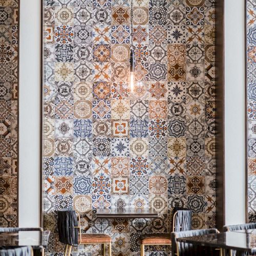 Decorative tiled archway wall with intricate blue, white, and earthy patterned tiles above a small dining area with dark wooden chairs and tables.