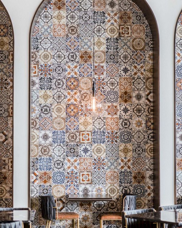 Decorative tiled archway wall with intricate blue, white, and earthy patterned tiles above a small dining area with dark wooden chairs and tables.
