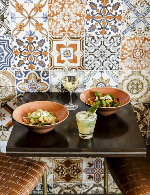 Two bowls of salad with a small green garnish, a lime-green drink, and a patterned tiled wall in the backdrop, set on a dark table.