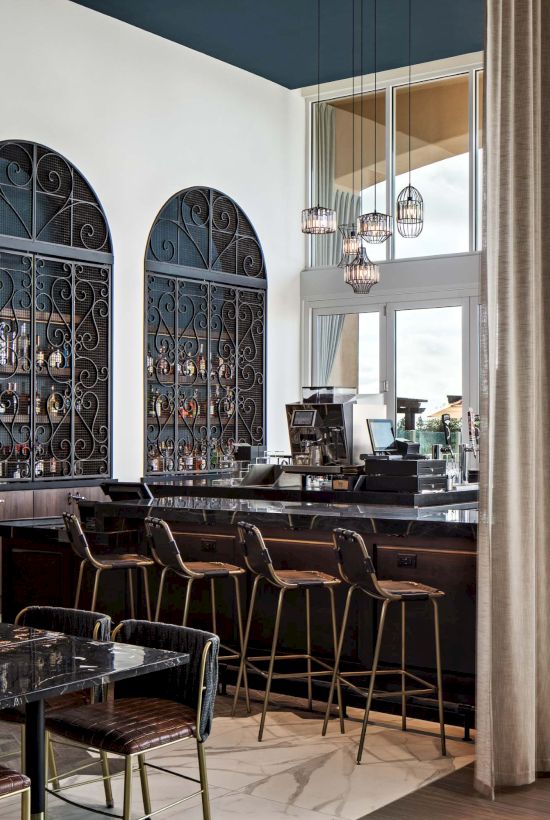 A stylish bar/restaurant interior with dark wood and arched alcoves, a long counter, high stools, and ample natural light from large windows.