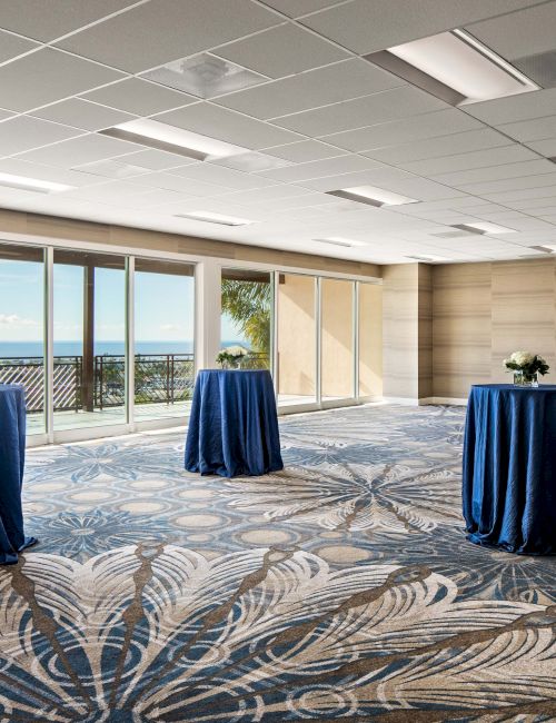 A bright event room with large windows showing a seaside view, and several tall round tables draped in blue cloths, ready for a gathering.