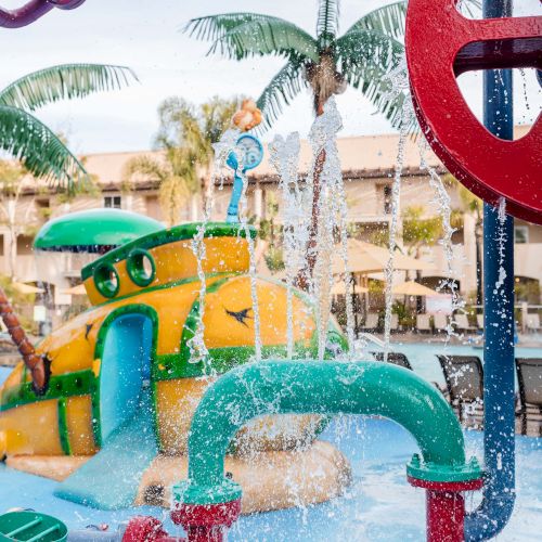 Kids splash and play on a colorful water park with slides, fountains, and palm trees under sunny skies.