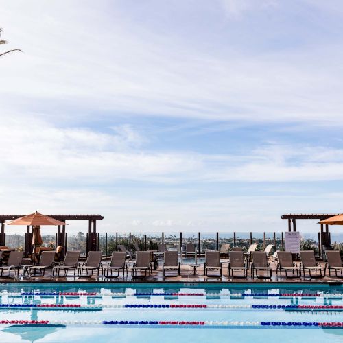 A sunny poolside scene with lounge chairs, umbrellas, palm trees, and a calm blue pool overlooking the ocean in the distance.