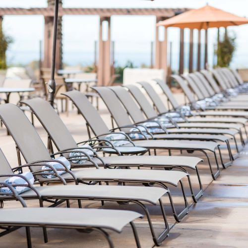 A row of empty lounge chairs on a sunny poolside, lined up under a bright, open deck with umbrellas and palm trees in the distance.