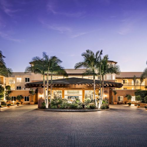 A luxurious resort courtyard at dusk with palm trees, warm yellow-lit buildings, and a central entry plaza, inviting and serene.