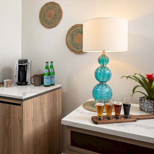 A modern hotel kitchenette with a coffee station, teal glass lamp, tropical plant, and a tray of four drinks on a white counter.