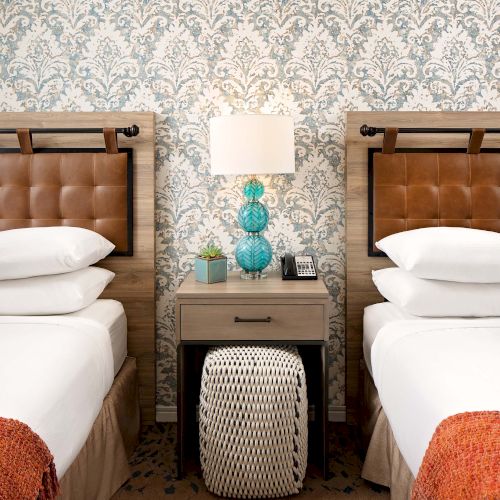 Two hotel beds with tufted headboards, a shared nightstand, a lamp, patterned wallpaper, and decorative pillows and throws.