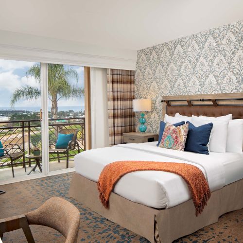A bright hotel room with a king bed, patterned wallpaper, teal lamps, and a balcony overlooking a pool and ocean, plus a desk and seating area.