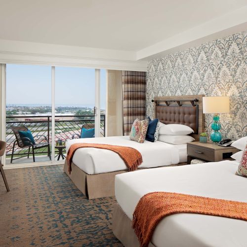 A stylish hotel room with two queen beds, patterned wallpaper, a desk, and a balcony with a city view and chairs, cozy lamps and colorful pillows.