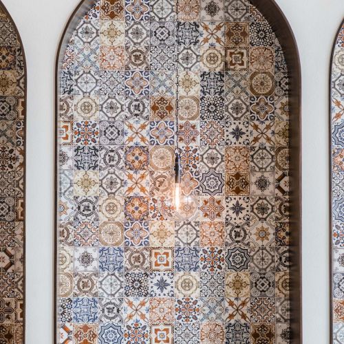 A tall arched wall features a colorful mosaic of geometric tile patterns in blue, beige, and brown, with small wooden chairs at the bottom.