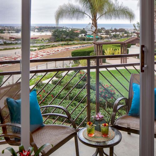 A cozy balcony with two chairs, a small table, blue cushions, and a view of a green lawn, palm trees, and distant houses by the coast, nice breeze.