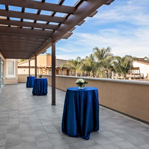 A sunny outdoor patio with a wooden pergola, potted plants, and a blue table covering on a tiled terrace overlooking a clear sky.