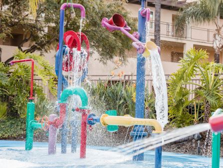 A colorful splash pad with water sprayers and fountains shooting water in multiple directions, surrounded by plants and a building in the background.