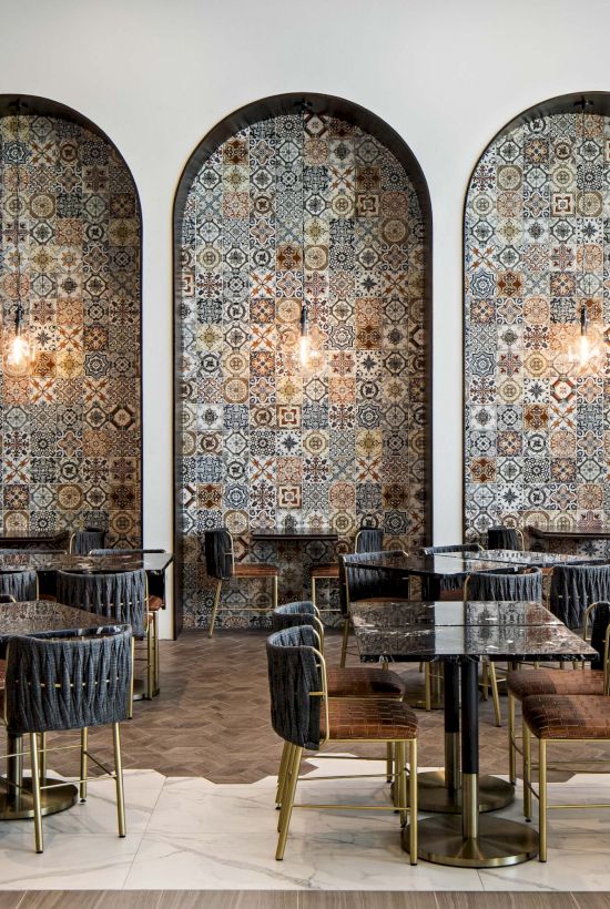 A stylish dining area with arched tiled backdrops, pendant lights, wood tables, and black/gold chairs in a chic restaurant setting.