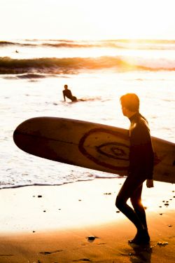 A person carrying a surfboard walks along a sunny beach at sunset, with waves and another surfer in the water, rocks nearby.